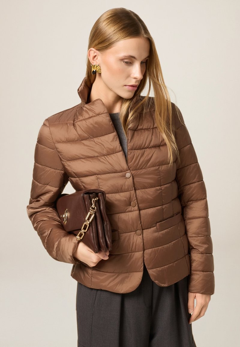 Braun gesteppte Jacke mit hohem Kragen, ausgestattet mit einem Knopfverschluss und Fronttaschen. Das Modell hält eine braune Tasche mit einer Kettenriemen.