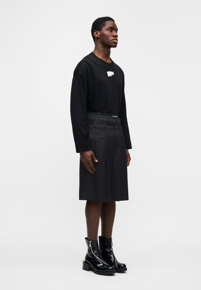 Μαύρο πουκάμισο με μακριά μανίκια συνδυασμένο με μαύρη φούστα μέχρι το γόνατο με υφασμένη μέση, φορεμένο με γυαλιστερά μαύρα μποτάκια αστραγάλου.