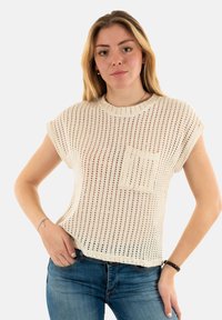 Crème-kleurige gebreide top met korte mouwen en een losse pasvorm, met een gestructureerde open weving en een voorzak. Gecombineerd met blauwe denim jeans.