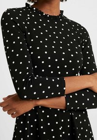 Musta mekko valkoisilla pilkuilla, vartalonmyötäinen yläosa, levenevä helma, pitkät hihat ja röyhelöinen pääntien yksityiskohta. Kankaan pinta on teksturoitu.