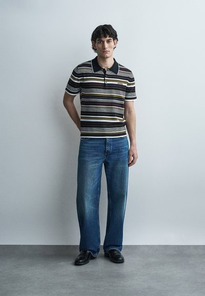 Νεαρός άνδρας στέκεται μπροστά σε έναν απλό τοίχο, φορώντας ριγέ πόλο μπλουζάκι, μπλε τζιν και μαύρα παπούτσια, με το ένα χέρι πίσω από την πλάτη του.