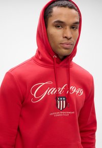 Jeune homme portant un sweat à capuche rouge avec "Gant 1949" et un blason de vêtements de sport américain sur la poitrine, regardant vers la caméra.