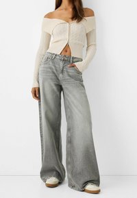 Krémový pletený top s odhalenými rameny a vzorem copánků, kombinovaný se světle šedými širokými džínami a béžovými teniskami s gumovou podrážkou.