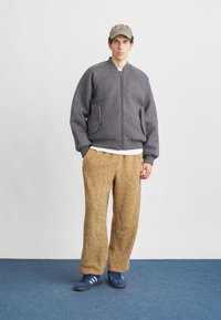 Chaqueta bomber de forro polar gris con cierre de cremallera, combinada con pantalones de pierna ancha marrones, texturizados y peludos. Zapatillas azules con suelas negras completan el look.