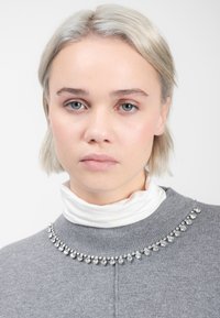 Grijze trui met een geribde halslijn, voorzien van een decoratieve rij kleine heldere rhinestones langs de kraag. Witte coltrui eronder.