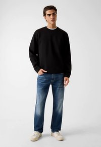 Schwarzer Baumwollpullover mit Rundhalsausschnitt, getragen über einem weißen Hemd, kombiniert mit hellblauen Jeans und cremefarbenen Sneakers.