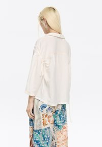 Light beige blouse with a collar, three-quarter sleeves, drawstring details, and subtle stitching, paired with colorful patterned trousers.