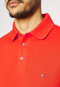 Man med kort skägg som bär en ljusröd pikétröja med två knappar knäppta och en liten flagglogga på bröstet.