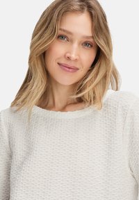 Femme souriante aux cheveux blonds portant un haut à manches longues texturé blanc sur fond clair uni.