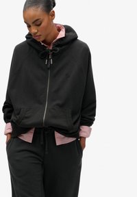 Felpa nera con zip e cappuccio regolabile, con tasche frontali, indossata sopra una camicia rosa con colletto; il tessuto è morbido e testurizzato, taglio oversize.