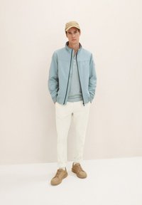 Giacca zip blu chiaro sopra una maglietta henley grigia, abbinata a pantaloni crema e sneakers marroni. Presenta una texture morbida e un design semplice.