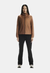 Femeie statând cu fața înainte, îmbrăcată cu o jachetă maro cu fermoar, pantaloni negri și adidași albi, pe un fundal alb.