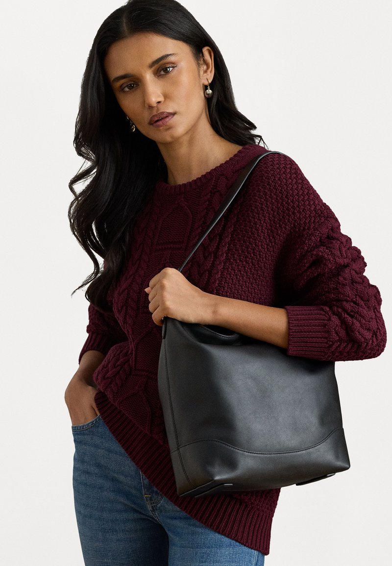 Μαύρη δερμάτινη τσάντα με ομαλή υφή και μια διακριτική καμπύλη βάση, κρατημένη από ένα άτομο που φοράει ένα μπορντό πλεκτό πουλόβερ και μπλε τζιν.