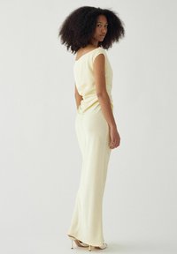 Femme aux cheveux bouclés naturels portant une robe longue jaune pâle sans manches et des sandales blanches à talons hauts, vue de profil.