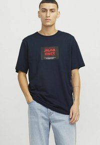 Námořnická modrá bavlněná tričko s krátkým rukávem a obdélníkovou grafickou nálepkou s nápisem "JACK & JONES" v červené a černé barvě. Pohodlný střih.