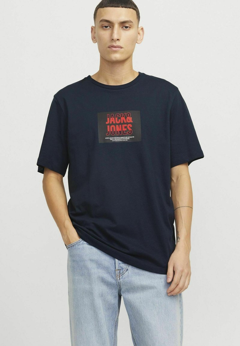 Námořnická modrá bavlněná tričko s krátkým rukávem a obdélníkovou grafickou nálepkou s nápisem "JACK & JONES" v červené a černé barvě. Pohodlný střih.