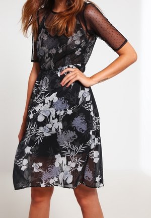 Vrouw gekleed in een zwarte doorschijnende jurk met bloemenpatronen en mouwen van gaas met polkadots, poserend met één hand op haar heup.