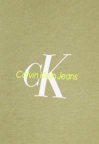Oliivinvihreä kangas, jossa on poimuttunut rakenne ja valkoinen "CK"-logo, ja sen alla keskitetty keltainen teksti "Calvin Klein Jeans".