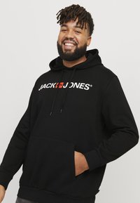 Sort hættejakke med en lomme foran og snore, med mærkenavnet "JACK & JONES" i fed hvid og rød skrift over brystet.