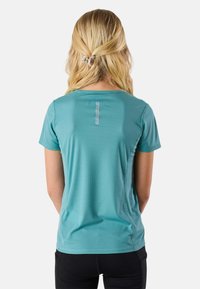 Lyshåret kvinde med hårklemme iført turkis kortærmet skjorte og sorte bukser, set bagfra mod en ensfarvet hvid baggrund.