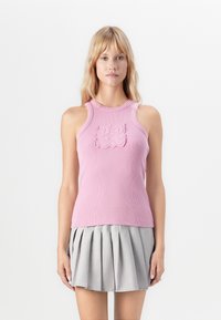 Woman wearing a ribbed pink sleeveless top with a textured logo and a gray pleated mini skirt, standing against a plain white background.
