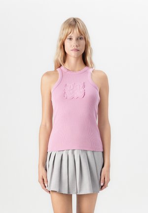 Woman wearing a ribbed pink sleeveless top with a textured logo and a gray pleated mini skirt, standing against a plain white background.