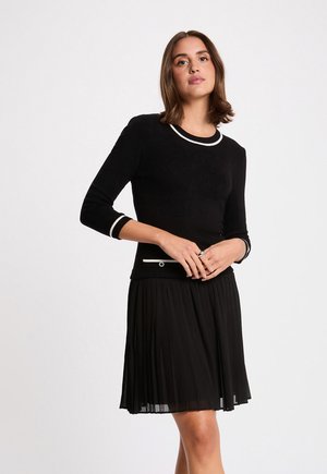 Mujer de pie con las manos entrelazadas, vistiendo un vestido negro con ribetes blancos en el escote, las mangas y el bolsillo, y una falda plisada.