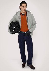 Szara bluza z zamkiem, pomarańczowa koszulka polo, granatowe spodnie, czarna torba i brązowe buty. Akcesoria to czarne rękawiczki i zielony pasek.