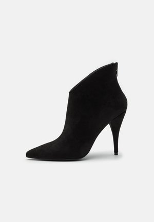 Bottine noire en suède à bout pointu, talon stiletto haut et fermeture éclair à l’arrière sur un fond clair uni.