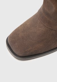 Braune Kunstleder-Ankle-Boots mit Quadratzehen, strukturiertem Obermaterial und kontrastierender schwarzer Gummisohle. Steppnähte entlang der Naht.