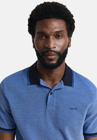 Modrá texturovaná polo košile s tmavým límcem a černými knoflíky. Obsahuje kontrastní modré pruhy na límci a rukávech. Logo značky na hrudi.