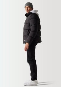 Chaqueta de plumas negra con diseño acolchado, cierre de cremallera y parche con el logo, llevada sobre una sudadera con capucha blanca y combinada con jeans negros y zapatos blancos.