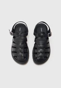 Par crnih kožnih sandala Calvin Klein s kopčama na trakama, tkanim dizajnom i unutarnjim potplatima s logom na svijetloj podlozi.