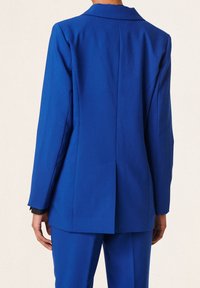 Osoba oblečená v jasně modrém přizpůsobeném sáčku a odpovídajících kalhotách, viděná zezadu na jednobarevném světlém pozadí.