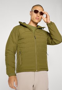 Olivenfarget polstret jakke med glidelås, justerbar hette og to sidelommer; brukt med beige solbriller mot en ensfarget bakgrunn.