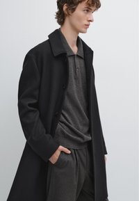 Zwarte wollen overjas met een reverskraag, gedragen over een gestructureerde grijze polo. Slim-fit grijze broek met krijtstreep. Minimalistisch ontwerp met knopen.