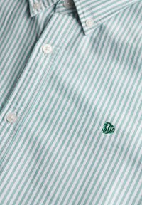 Camisa de botones de rayas azul claro y blanca. Presenta botones blancos, un collar y un logo bordado en verde en el pecho. Textura de tela suave.