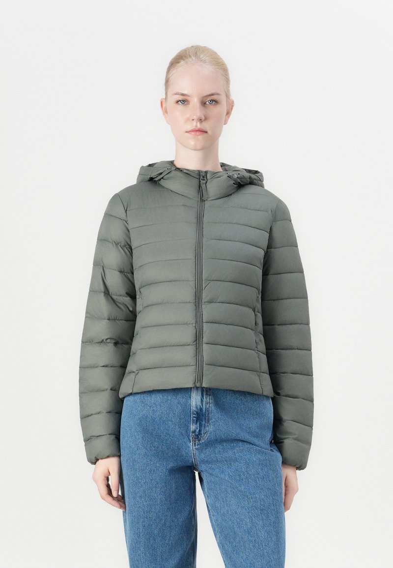 Polstret olivengrønn jakke med hette og glidelås foran, med horisontalt quiltede seksjoner og en kort design. Kombinert med blå jeans.