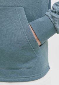 Sweatshirt bleu clair avec poche kangourou, poignets côtelés et texture douce, avec des coutures inclinées à l'ourlet et aux manches.