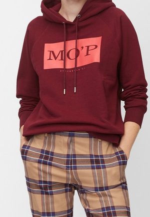 Person iført en vinrød hættetrøje med teksten "MO’P" og beige ternede bukser, hænderne i lommerne, mod en ensfarvet lys baggrund.