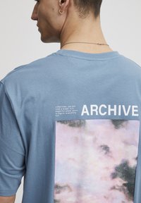 Sinine puuvillane t-särk, mille seljal on graafiline print pilvedest, koos sõnaga "ARCHIVE" ja valge tindiga tekstiga.