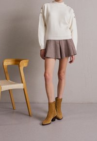 Witte geribde trui met gouden knoopaccenten, gecombineerd met een bruine geruite rok. Model draagt tan suède enkellaarsjes met een dikke hak.