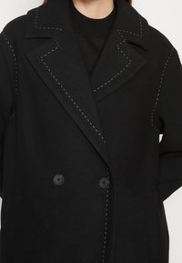 Cappotto nero doppiopetto con cuciture bianche a contrasto, rever oversize, tessuto testurizzato e due bottoni.