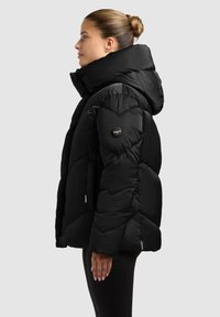 Schwarze Daunenjacke mit hohem Kragen und Kapuze, mit gesteppter Textur, seitlichen Taschen und einem Logopatch am linken Ärmel.