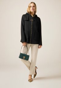 Abrigo negro con capucha y cremallera dorada, acompañado de pantalones crema, un bolso verde y tacones estampados.