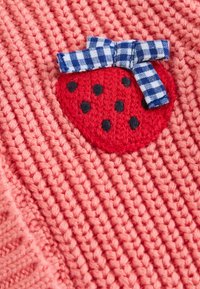 Adīta korāļkrāsas auduma ar reljefainu rakstu rotā sarkans zemeņu aplikācijas uzšuvums, ko papildina melni punkti un zilbalts rūtainas lentes loks.