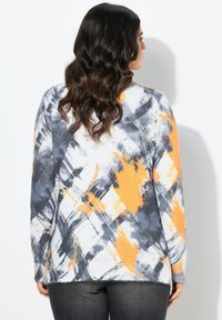 Eine Frau mit langen, welligen, dunklen Haaren trägt einen bunten Pullover mit abstraktem Muster in Grau-, Weiß- und Orangetönen sowie eine dunkle Hose, während sie vor einer weißen Wand steht.