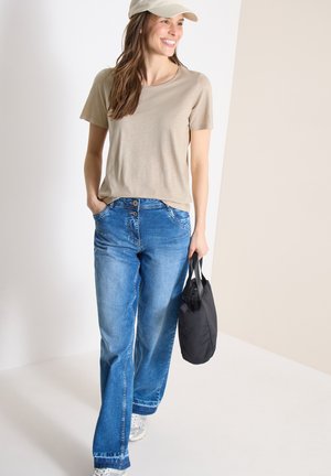 Glimlachende vrouw met een beige pet, beige t-shirt, blauwe wijd uitlopende jeans, witte sneakers, een zwarte handtas dragend, lopend in een neutrale binnenruimte.