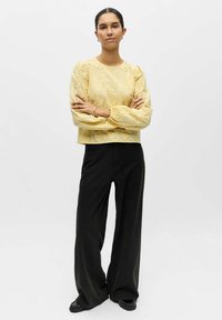 Haut jaune à manches longues brodé avec un col rond et des manches froncées, associé à un pantalon noir à jambes larges taille haute et des sandales.