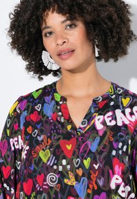 Kleurvolle blouse met patroon in zwarte stof, versierd met harten en woorden als "VREDE", met een v-hals en een subtiele glans. Witte statement oorbellen.